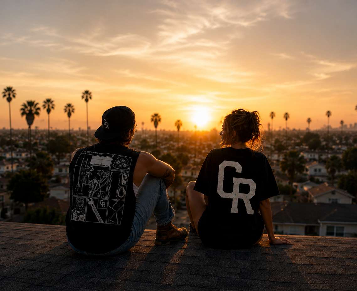 Two people wearing Cinder Route t-shirts sitting on a rooftop at sunset overlooking palm trees